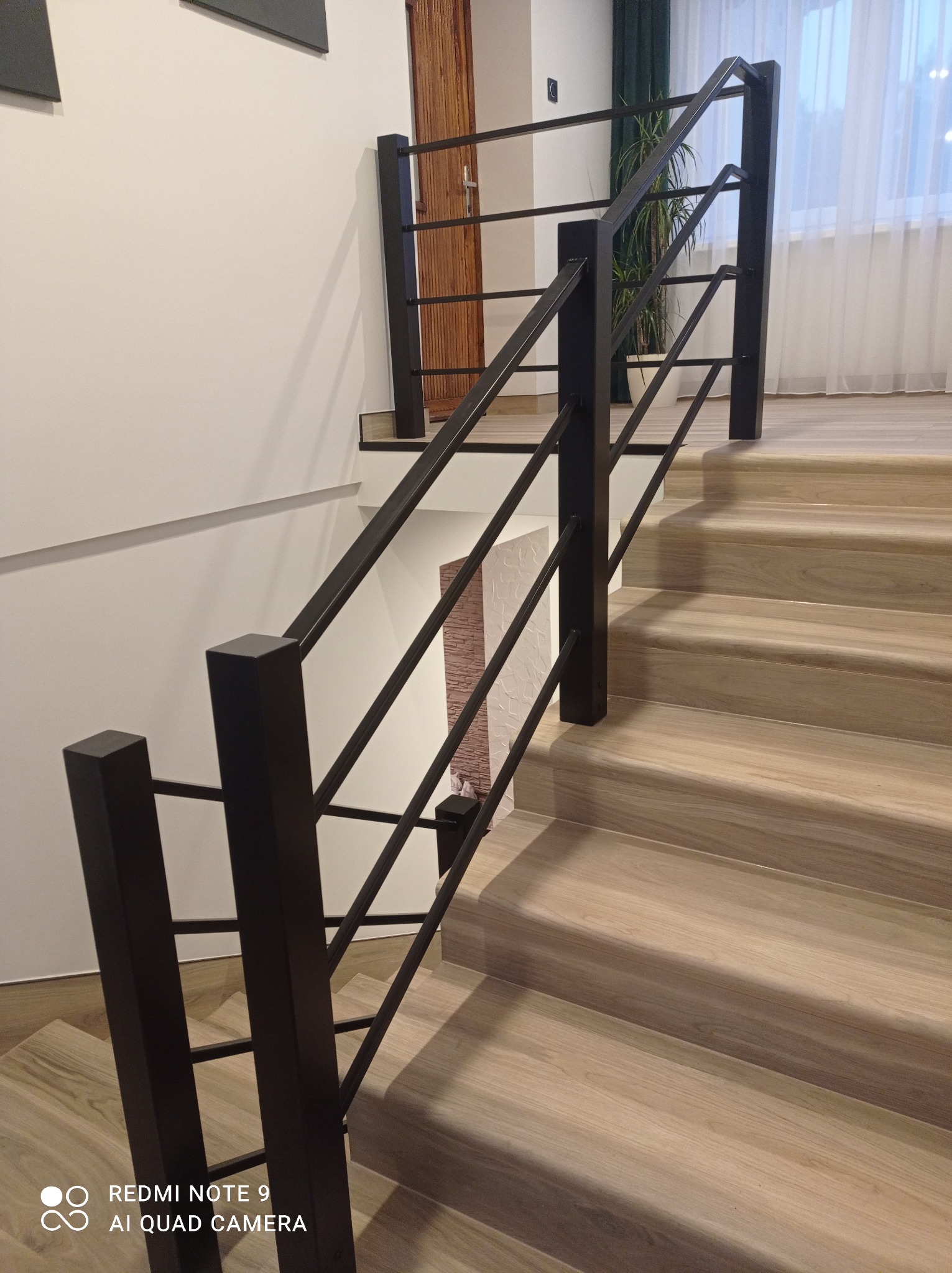 Nowoczesna klatka schodowa z drewnianymi stopniami i czarną, minimalistyczną balustradą z metalowymi prętami, widok z dołu schodów.