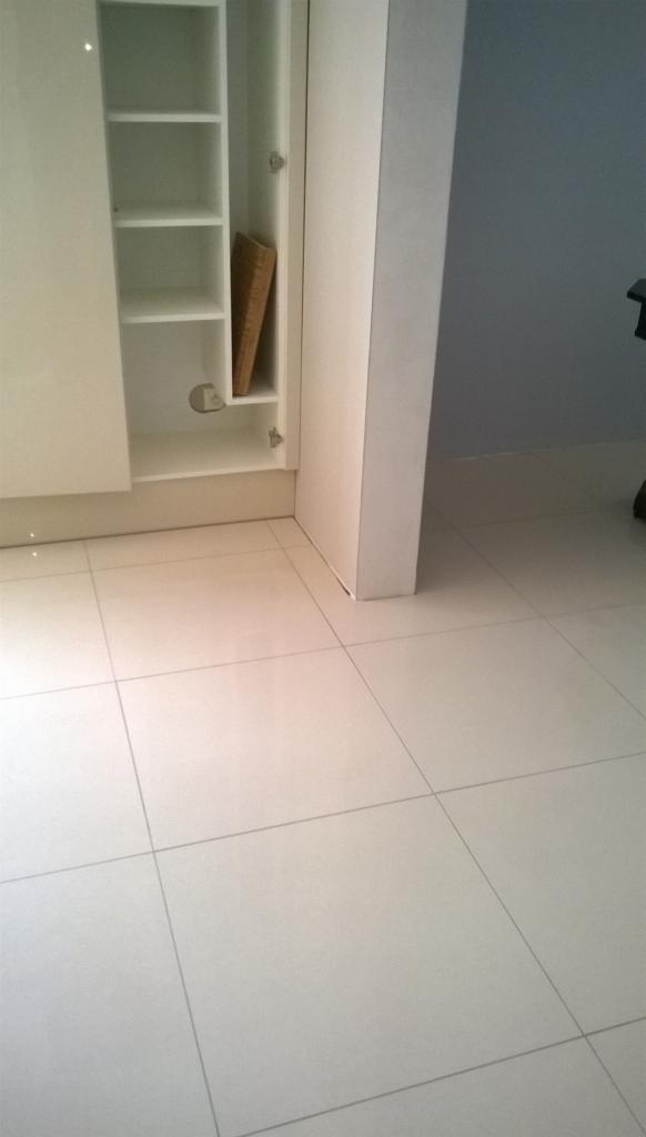 Jasne, kwadratowe płytki podłogowe z wąskimi fugami, fragment białej szafki z półkami i widocznym otworem na rurę oraz fragment ściany pomalowanej na szaro.