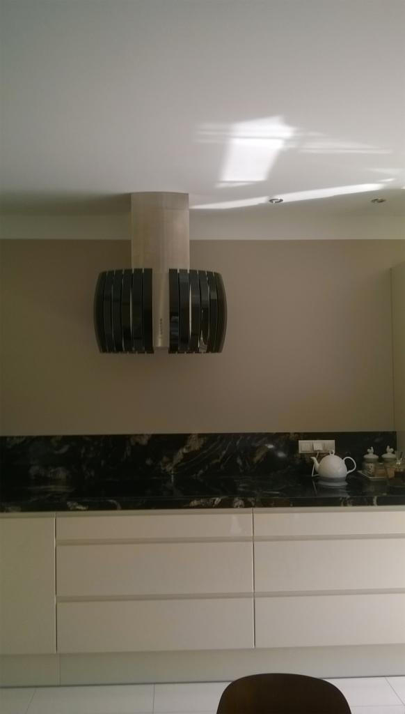 Nowoczesny okap kuchenny z czarnymi akcentami, zamontowany nad blatem z czarnego marmuru, uzupełniony białymi szafkami i czajnikiem.