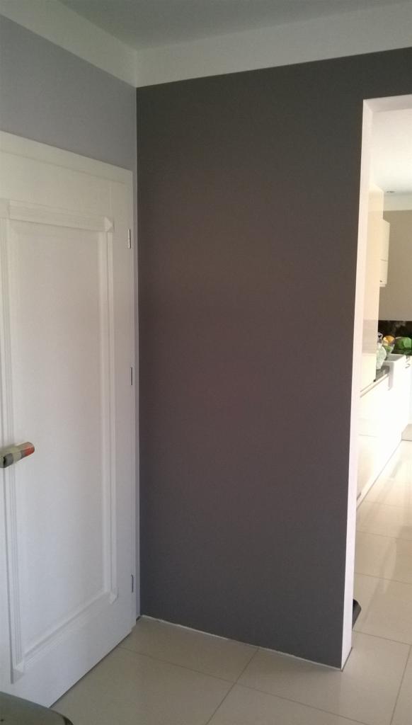 Pomieszczenie po malowaniu: szara ściana sąsiaduje z białą ścianą i drzwiami, widoczna podłoga z płytek i fragment kuchni w tle.