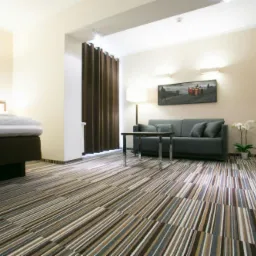 Pokój hotelowy z wzorzystą wykładziną w paski, szarą sofą i łóżkiem. Widoczne oświetlenie ścienne i dekoracje. Ciemne zasłony w tle dodają przytulności.