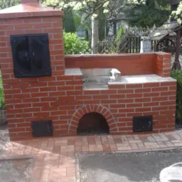 Murowany grill ogrodowy z czerwonej cegły, z czarnymi, metalowymi drzwiczkami, usytuowany na wyłożonym cegłą patio, w tle zieleń ogrodu.