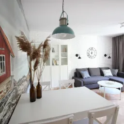 Jasne wnętrze pokoju z dużym stołem, szarą sofą i fototapetą z motywem czerwonego domku na ścianie.