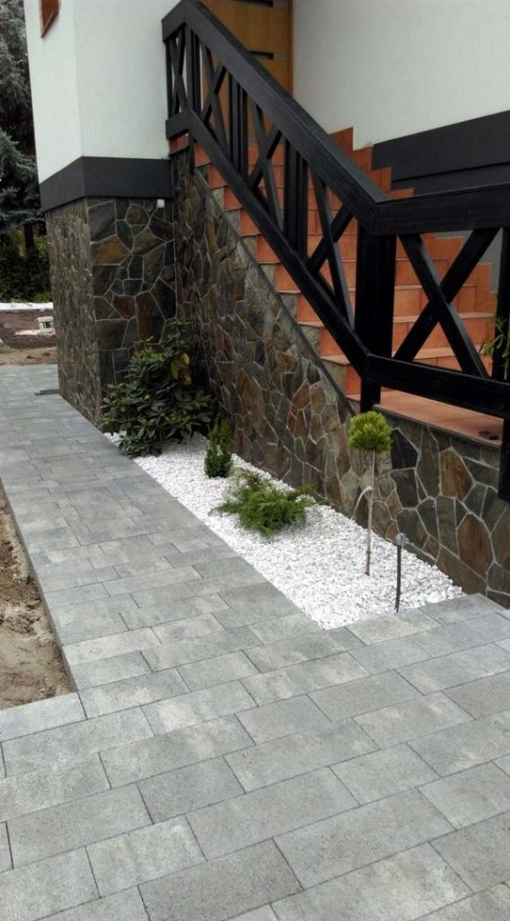 Ułożona szara kostka brukowa prowadząca do schodów z czarną balustradą i kamienną okładziną, obok rabata z białym grysem i niskimi roślinami.