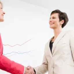 Dwie uśmiechnięte kobiety w biurowych strojach ściskają sobie dłonie na tle białej ściany z wykresem wzrostu. Jasne oświetlenie, biznesowa atmosfera.