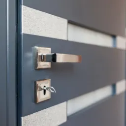 Szare drzwi wewnętrzne z poziomymi, dekoracyjnymi wstawkami, chromowaną klamką i kluczem w zamku.