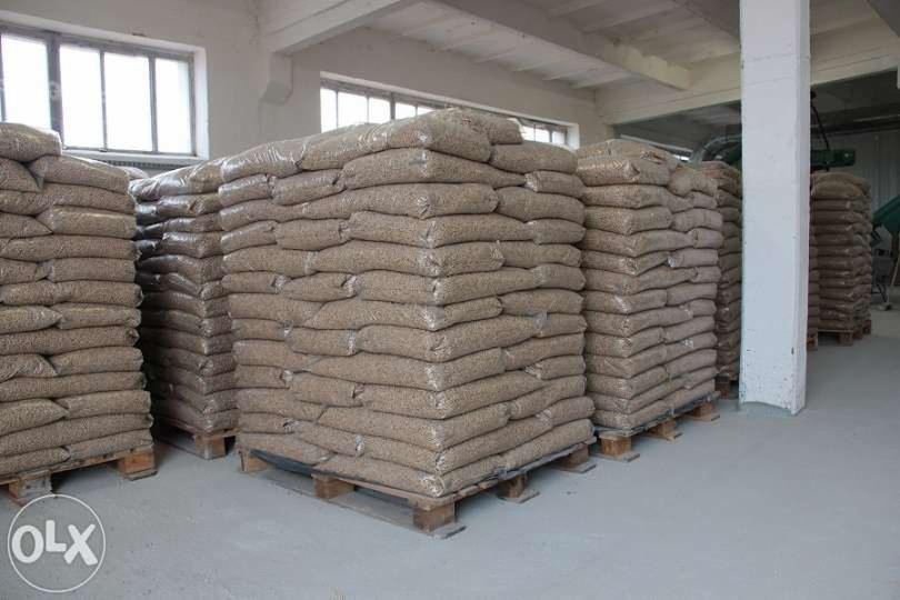 Stosy worków z pelletem na paletach w magazynie. Widoczne okna i betonowy filar. Pellet w przezroczystych workach, ułożony równo, gotowy do transportu.