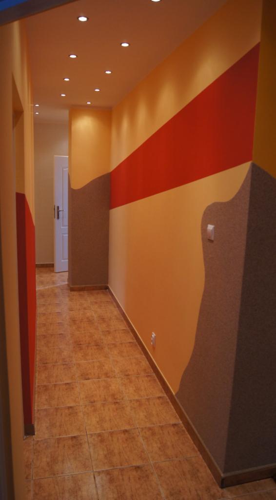 Korytarz po malowaniu z dekoracyjnymi pasami w odcieniach pomarańczy, brązu i czerwieni. Podwieszany sufit z punktowym oświetleniem. Na podłodze brązowe płytki.