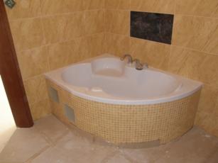 Narożna, biała wanna obudowana mozaiką w łazience z beżowymi płytkami. Widoczne baterie i fragment drewnianej futryny. Ujęcie z góry, lekko z prawej strony.