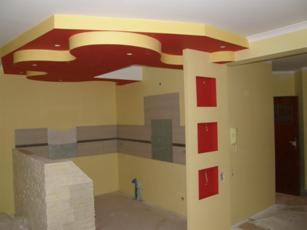 Żółto-czerwony, wielopoziomowy sufit podwieszany z dekoracyjnymi elementami, częściowo wykończona ściana z płytkami w pomieszczeniu w trakcie remontu.