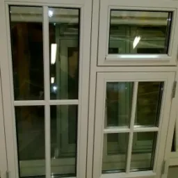 Białe, czteroskrzydłowe okno z szybami zespolonymi i szprosami, widoczne zawiasy. W szybach odbija się wnętrze pomieszczenia.