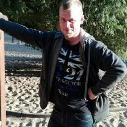 Mężczyzna w dżinsach i bluzie z kapturem opiera się o drewniany słupek na piaszczystym placu zabaw, w tle zieleń drzew. Uśmiech na twarzy, swobodna poza.