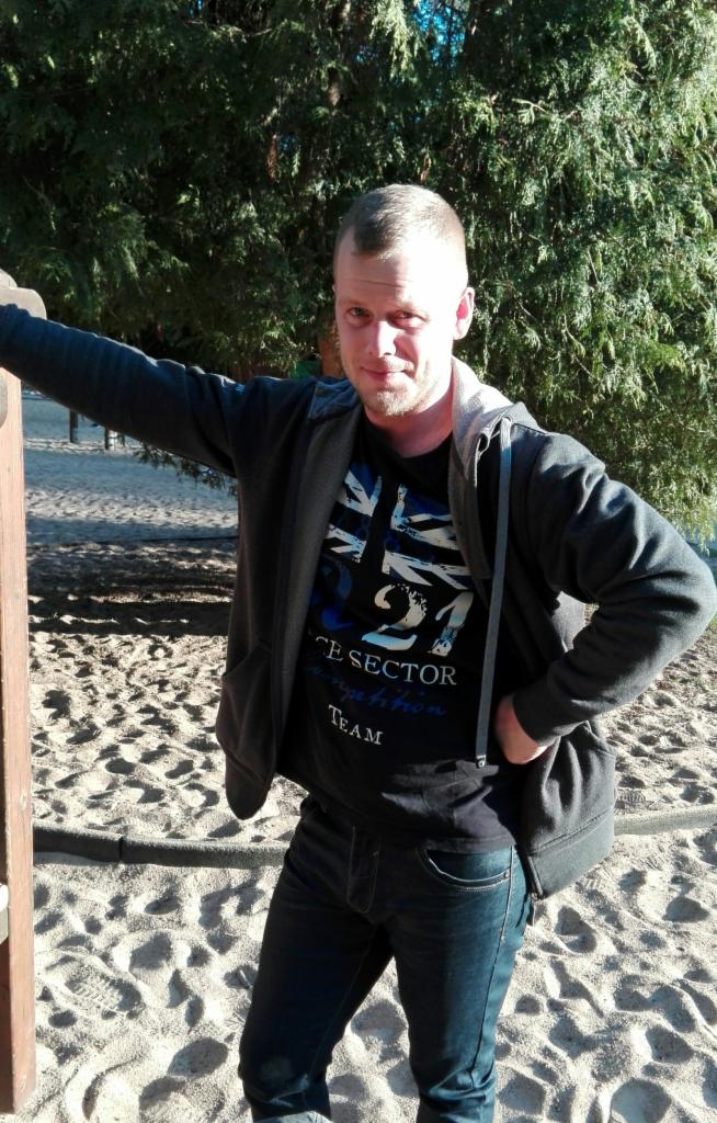 Mężczyzna w dżinsach i bluzie z kapturem opiera się o drewniany słupek na piaszczystym placu zabaw, w tle zieleń drzew. Uśmiech na twarzy, swobodna poza.