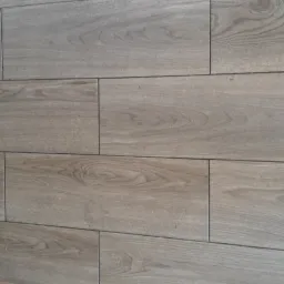 Ceramiczne płytki podłogowe imitujące drewno, ułożone w regularny wzór cegiełki, z widoczną przypodłogową listwą w kolorze zbliżonym do płytek.