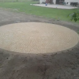 Okrągła powierzchnia z kostki brukowej ułożona w koncentryczny wzór na zewnątrz, z widocznym trawnikiem i budynkiem w tle.