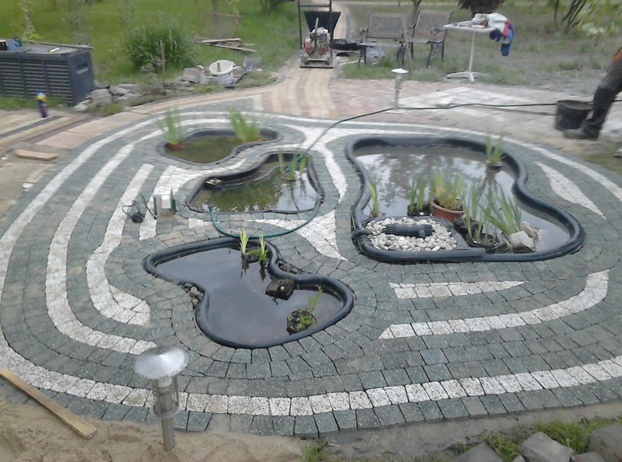 Ogród z oczkami wodnymi otoczonymi kostką brukową w geometryczny wzór, z roślinami wodnymi. Widoczne lampy ogrodowe i narzędzia budowlane.