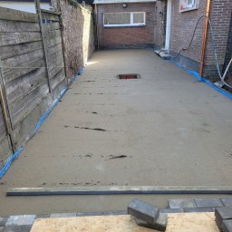 JANKO POSADZKI Janusz Dąbrowski - Świeżo wylana, jeszcze mokra, szara posadzka jastrychowa w wąskim przejściu między budynkiem a drewnianym płotem, zabezpieczona niebieską folią przy ścianach, z widocznym otworem rewizyjnym...