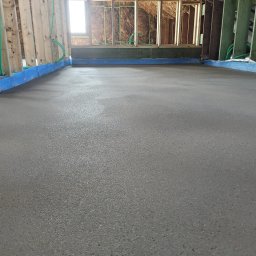 JANKO POSADZKI Janusz Dąbrowski - Świeżo wylana, gładka posadzka w surowym wnętrzu poddasza z widoczną konstrukcją drewnianą, izolacją i oknem.