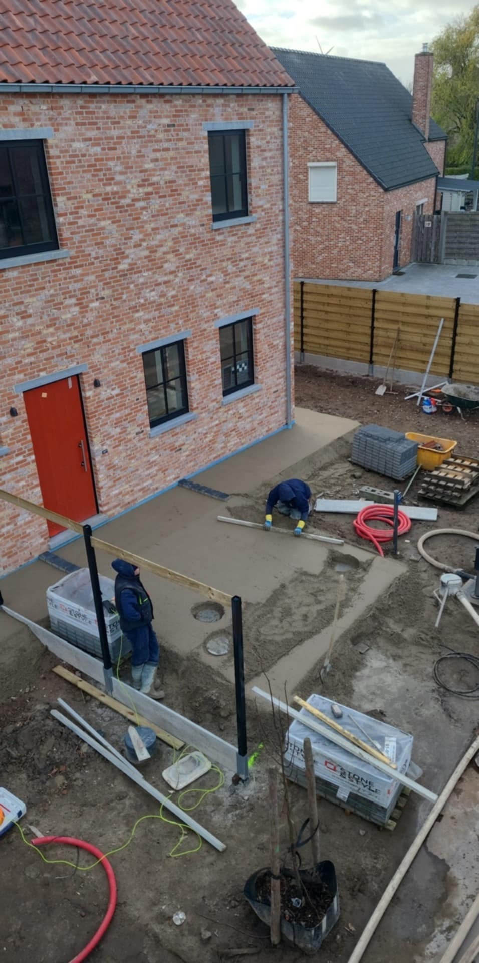 Wylewka jastrychowa na zewnątrz budynku z czerwonej cegły, dwóch pracowników w niebieskich kombinezonach wygładza powierzchnię, widoczne narzędzia, szalunki i przygotowane materiały budowlane.