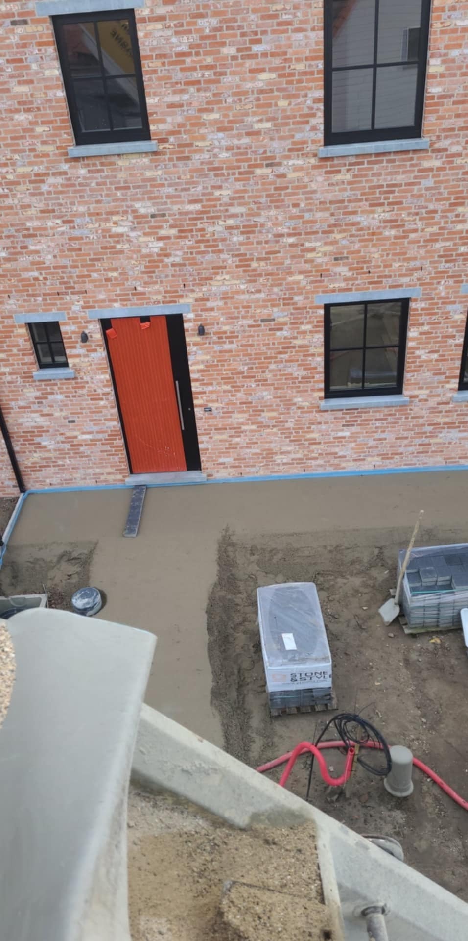 Świeżo wylana betonowa posadzka przed budynkiem z czerwonej cegły z pomarańczowymi drzwiami, widoczne narzędzia i materiały budowlane.