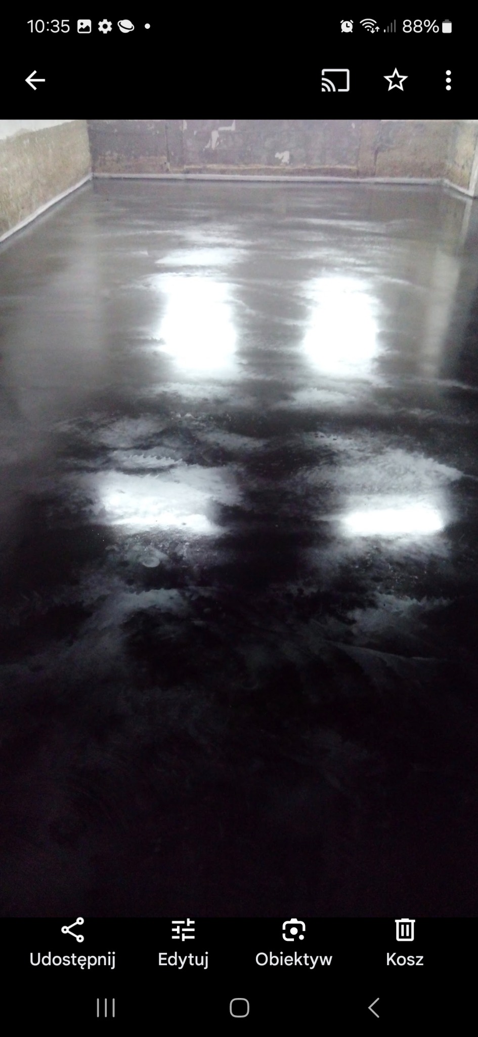 Gładka, ciemna posadzka w pomieszczeniu, z widocznymi refleksami światła na powierzchni, przypominająca garaż.