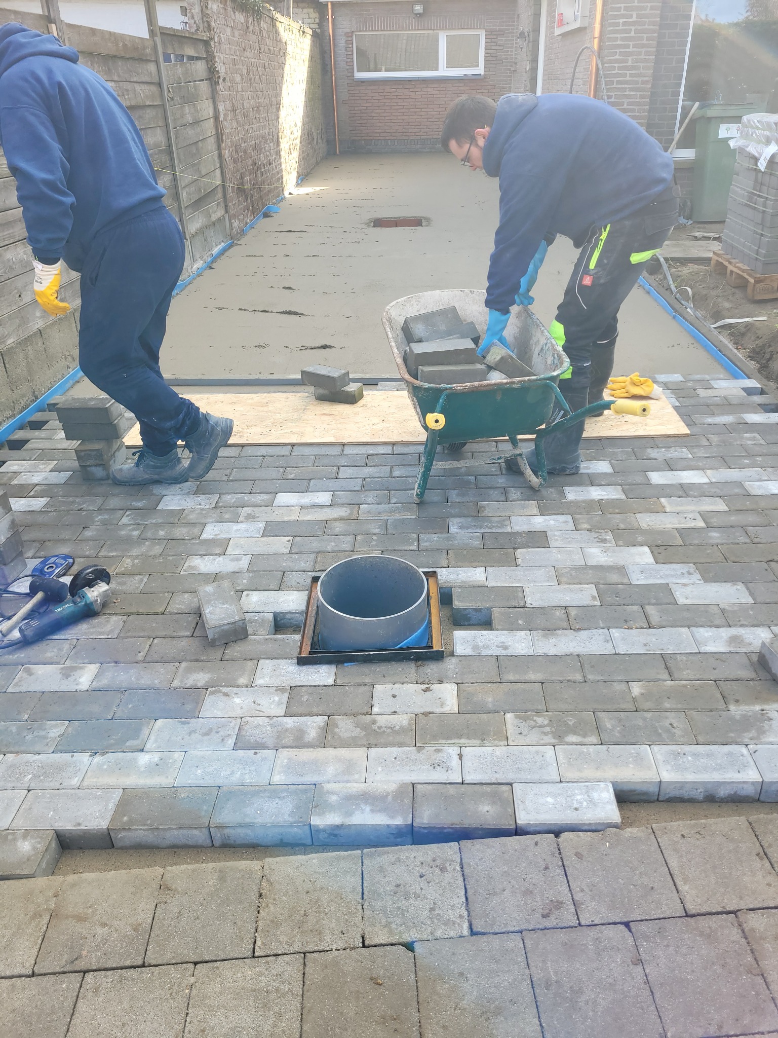 Układanie szarej kostki brukowej na podwórku: pracownik w niebieskiej bluzie i okularach wyjmuje kostkę z taczki, drugi pracownik stoi obok. Widoczne narzędzia i rury kanalizacyjne.