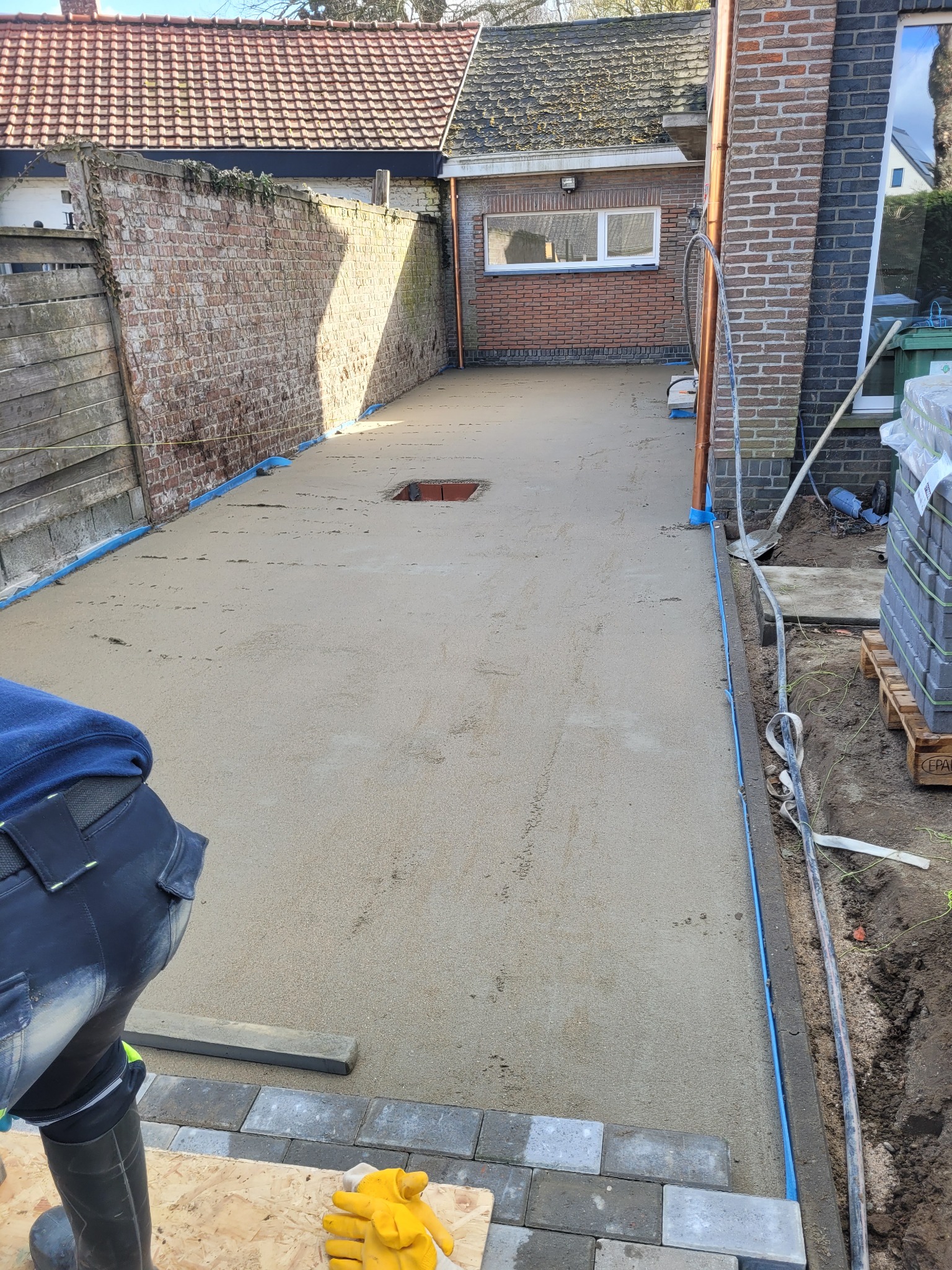 Świeżo wylana, szara posadzka z widocznym otworem rewizyjnym, zabezpieczona niebieską folią przy ścianach. W tle ceglany budynek i drewniany płot. W kadrze fragment pracownika układającego kostkę...