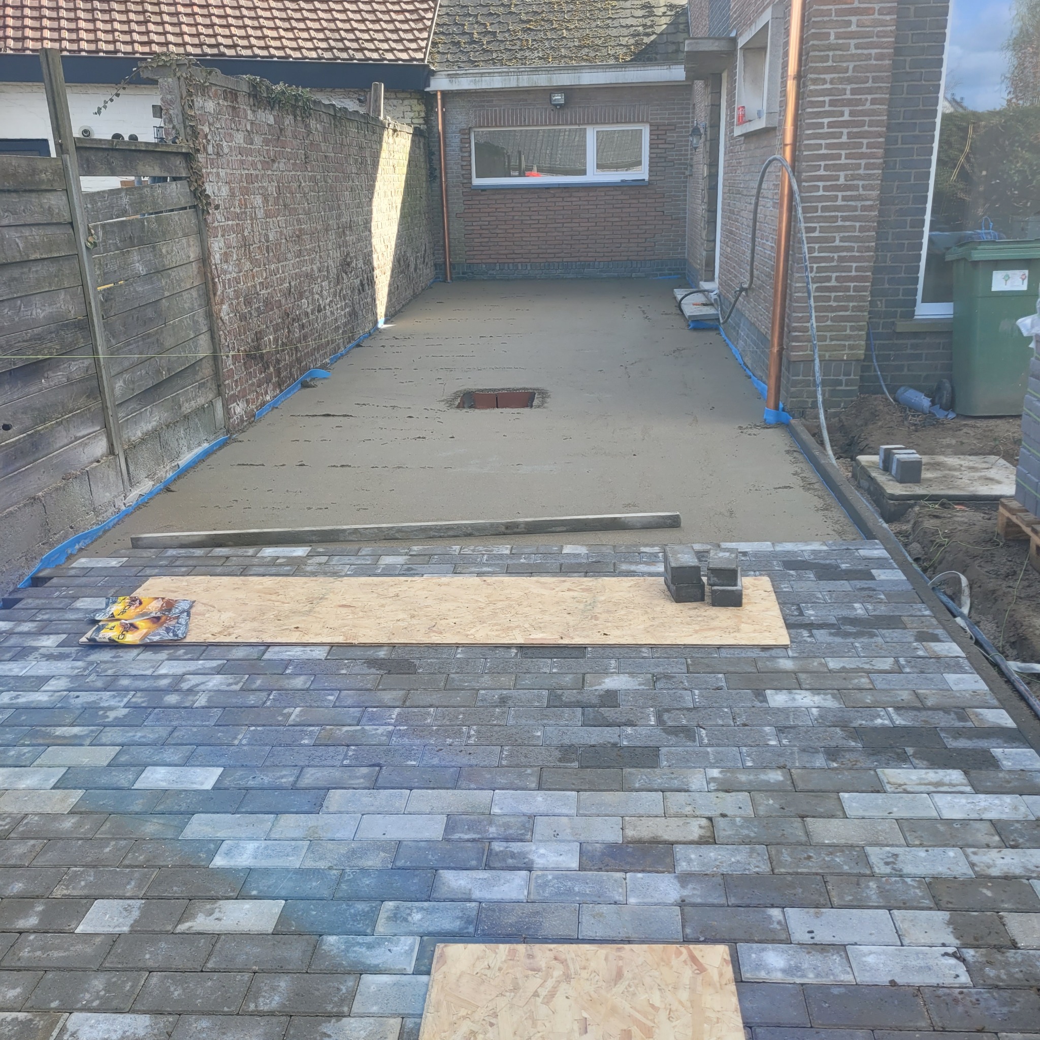 Przygotowanie podłoża pod kostkę brukową: Wylany jastrych z otworem rewizyjnym, częściowo ułożona kostka brukowa, deska i paczka kostki na ułożonej części.