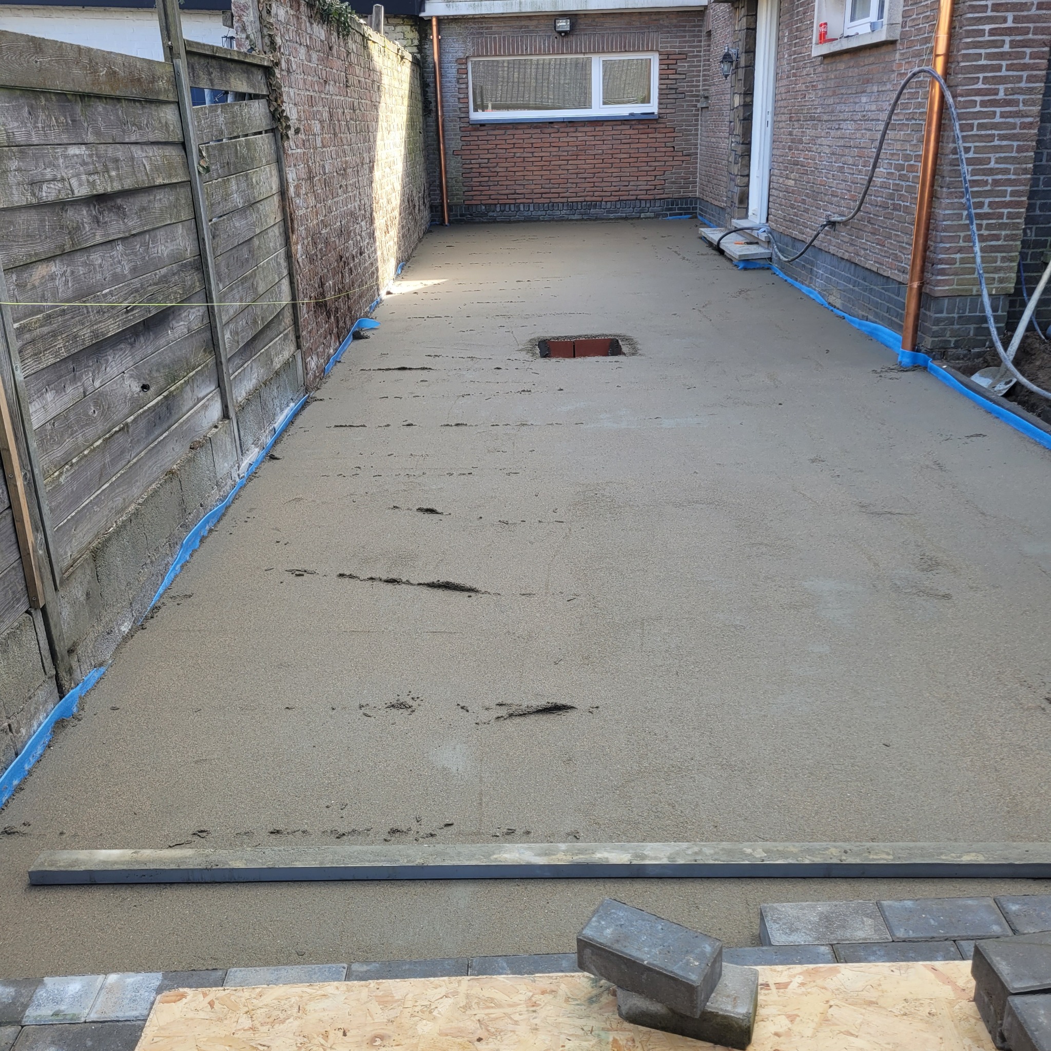 Świeżo wylana, jeszcze mokra, szara posadzka jastrychowa w wąskim przejściu między budynkiem a drewnianym płotem, zabezpieczona niebieską folią przy ścianach, z widocznym otworem rewizyjnym...