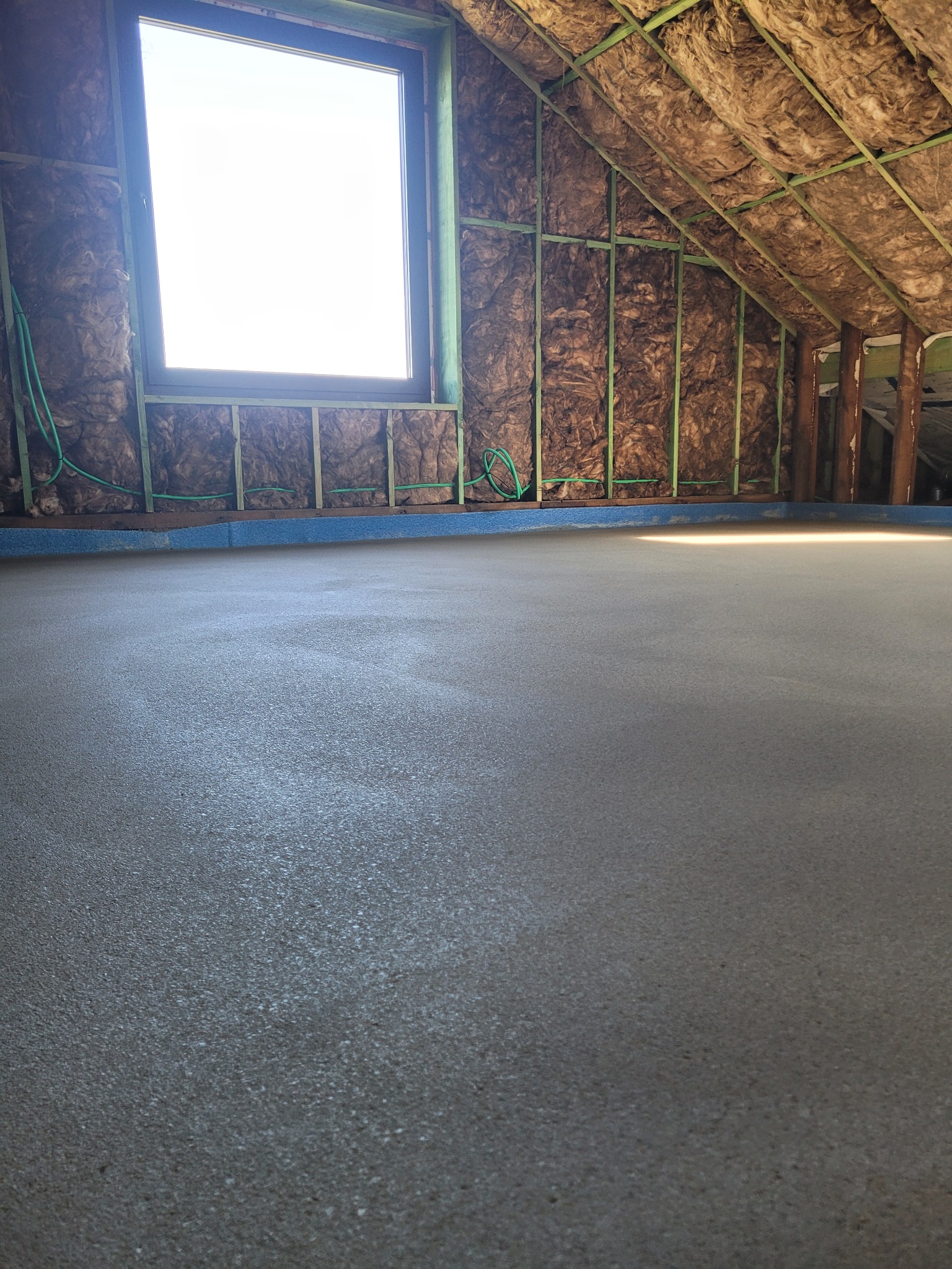 Świeżo wylana, gładka posadzka jastrychowa w poddaszu z oknem, izolacją z wełny mineralnej i elementami konstrukcji drewnianej.