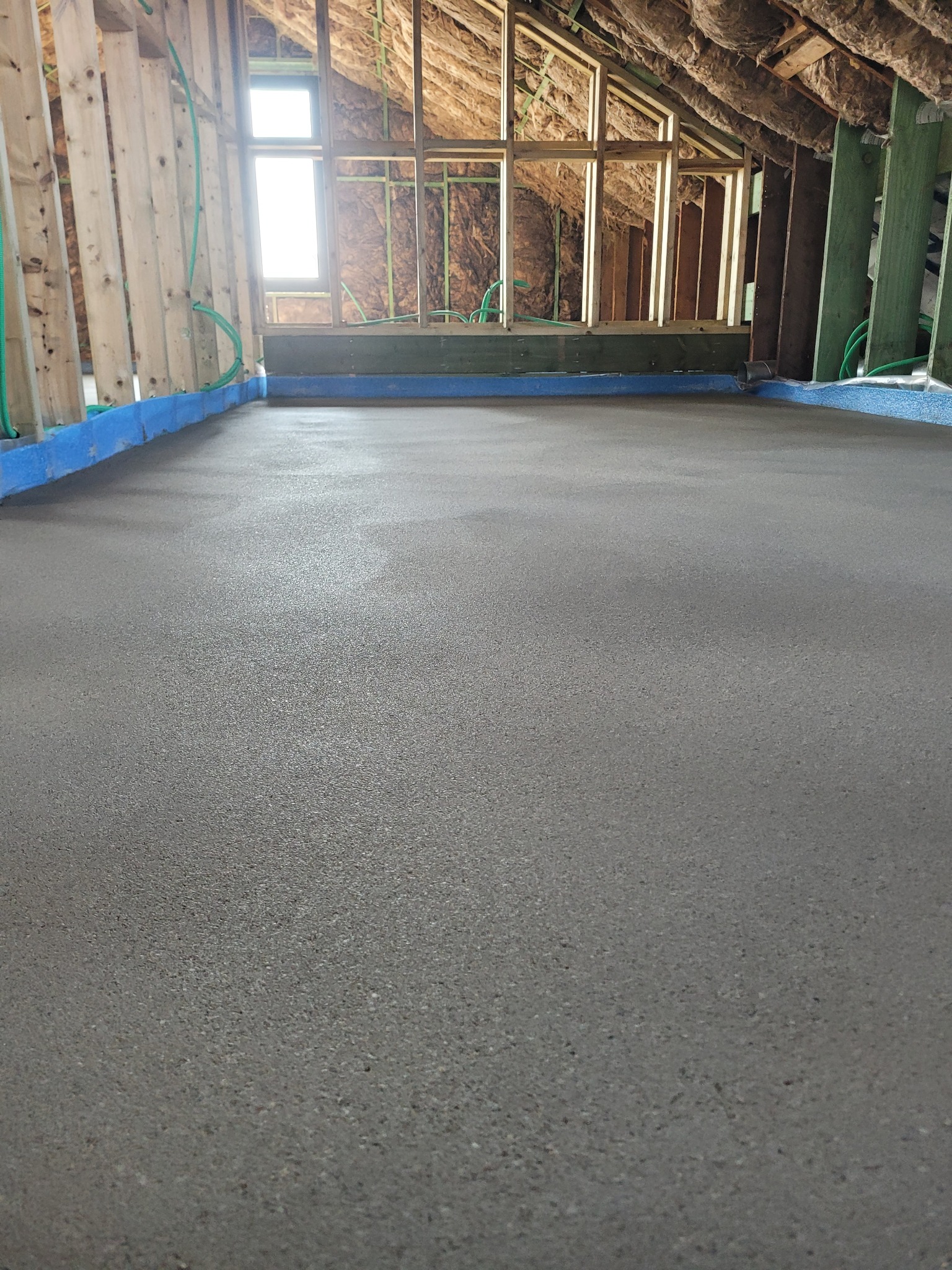 Świeżo wylana, gładka posadzka w surowym wnętrzu poddasza z widoczną konstrukcją drewnianą, izolacją i oknem.