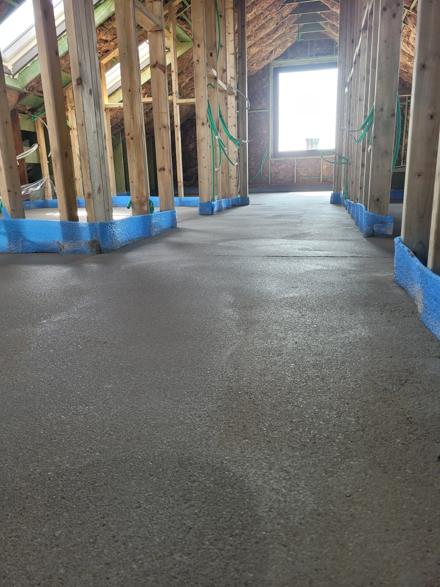 Świeżo wylana, mokra posadzka w surowym stanie, zabezpieczona niebieską taśmą przy drewnianych słupach konstrukcyjnych poddasza.