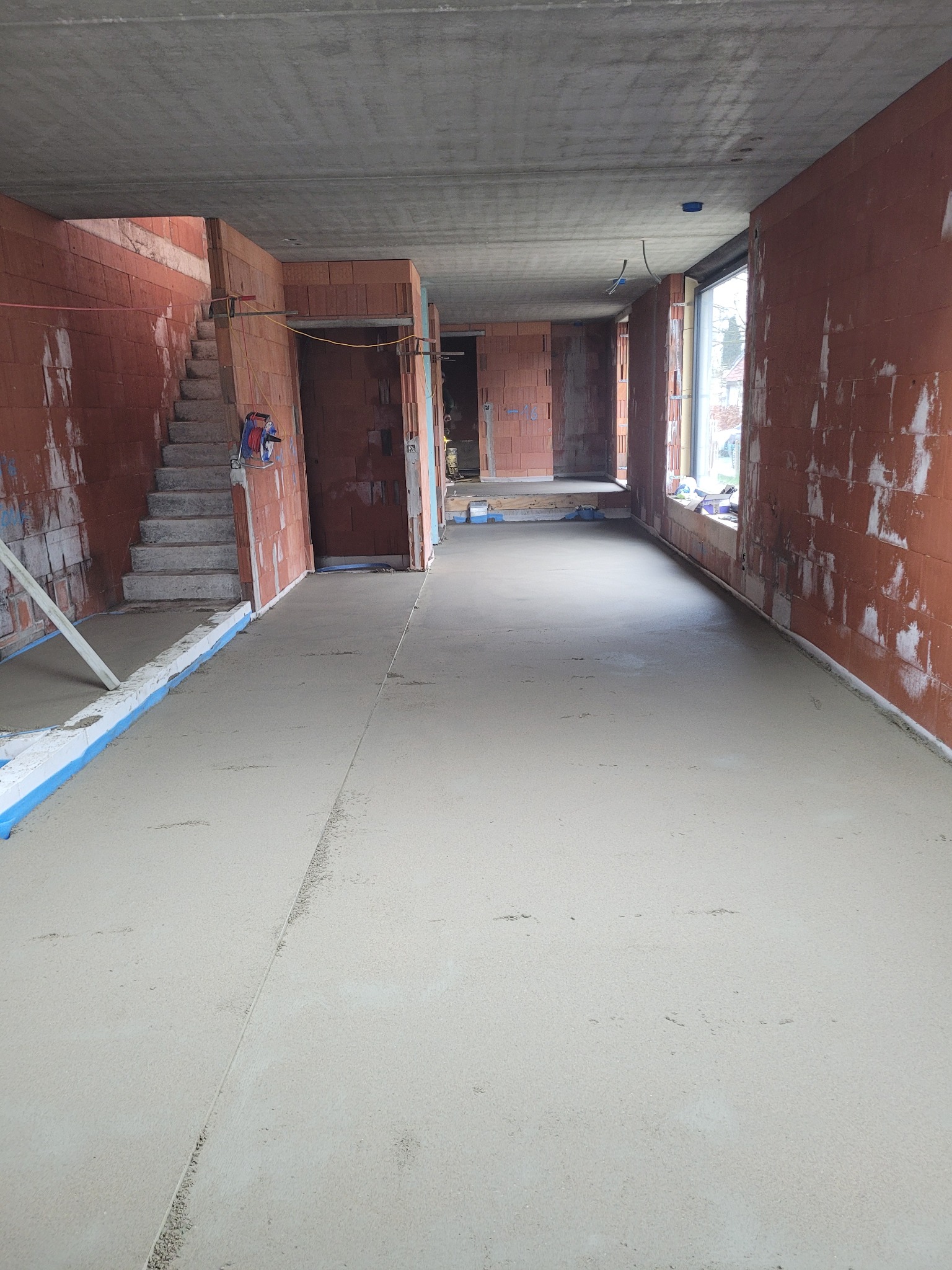 Świeżo wylana, jasnoszara posadzka w surowym, ceglanym wnętrzu budynku w trakcie budowy, widoczne schody i okno.