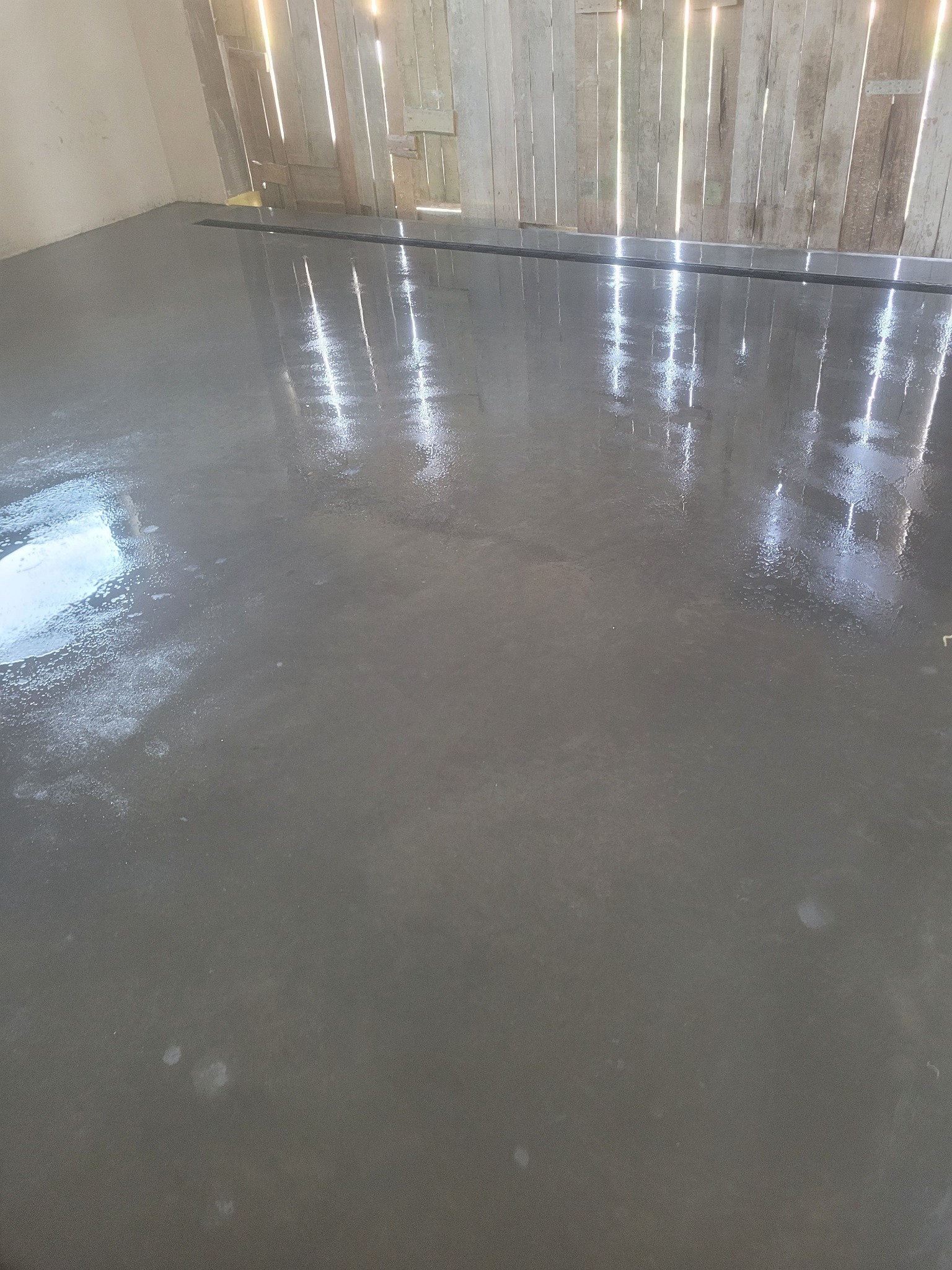 Świeżo wylana, gładka posadzka jastrychowa w pomieszczeniu z drewnianą ścianą, odbijająca światło. Widoczny odpływ liniowy w podłodze.