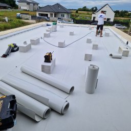 Cadart P.K.B. Krzysztof Piotrowski - Białe membrany dachowe w rolkach na płaskim dachu z bloczkami i narzędziami. W tle domy jednorodzinne i pracownik w koszulce MIG-BUD.