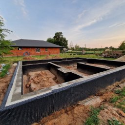 Cadart P.K.B. Krzysztof Piotrowski - Fundamenty domu jednorodzinnego z izolacją, wykonane w Raszynie. Widok na budynek w tle, teren budowy w naturalnym oświetleniu, z perspektywy lekko z góry.