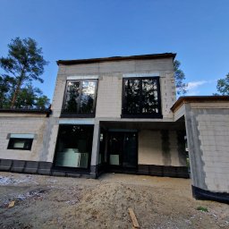 Cadart P.K.B. Krzysztof Piotrowski - Surowy stan nowoczesnego domu z betonu komórkowego, z dużymi oknami i widoczną konstrukcją, otoczonego drzewami. W tle błękitne niebo.