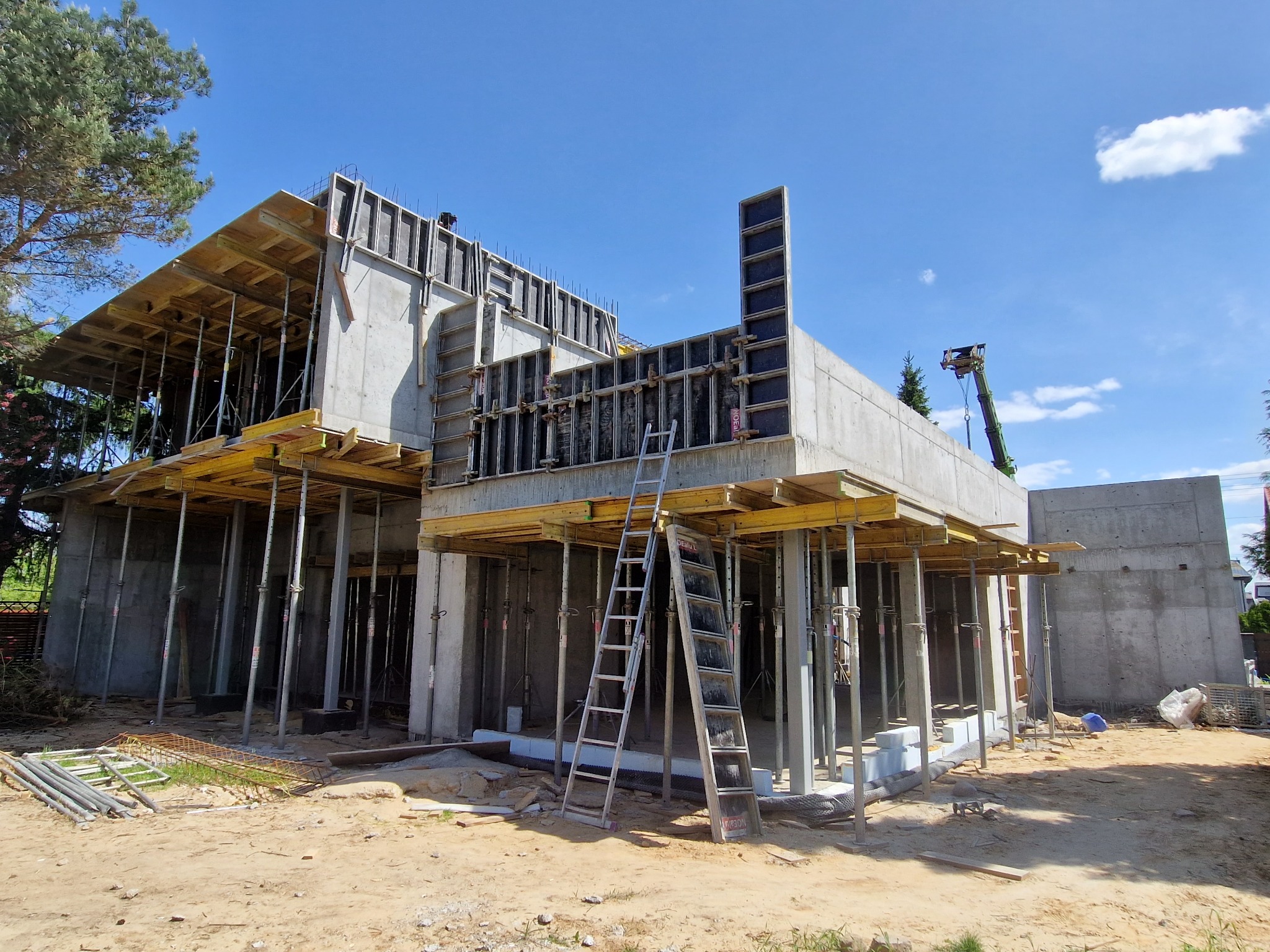 Surowy stan budynku w trakcie budowy z betonu i drewnianych szalunków, drabina oparta o ścianę, błękitne niebo w tle. Widoczne zbrojenia i podpory.