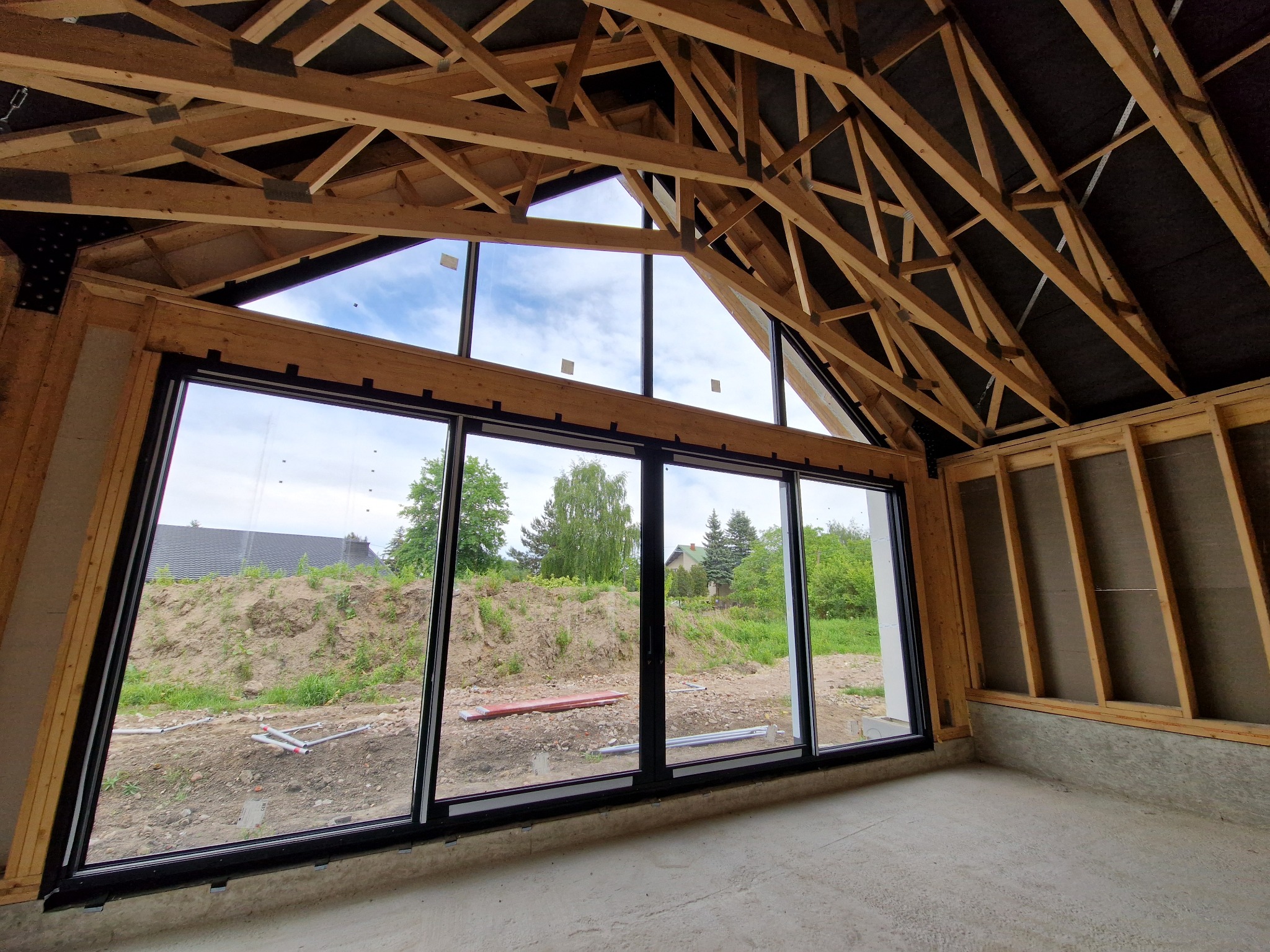 Wnętrze w stanie surowym otwartym z drewnianą konstrukcją dachu i dużymi przesuwnymi oknami z widokiem na zewnątrz. Widoczny etap budowy z elementami wykończeniowymi.