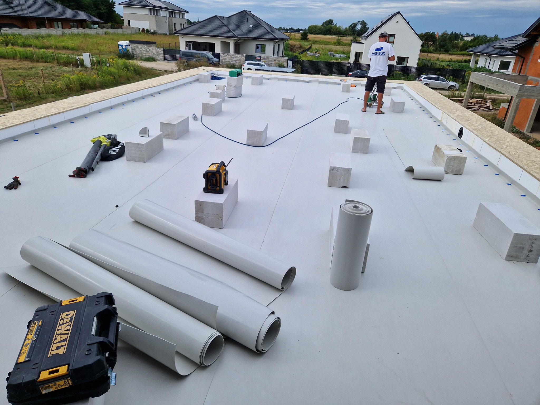 Białe membrany dachowe w rolkach na płaskim dachu z bloczkami i narzędziami. W tle domy jednorodzinne i pracownik w koszulce MIG-BUD.