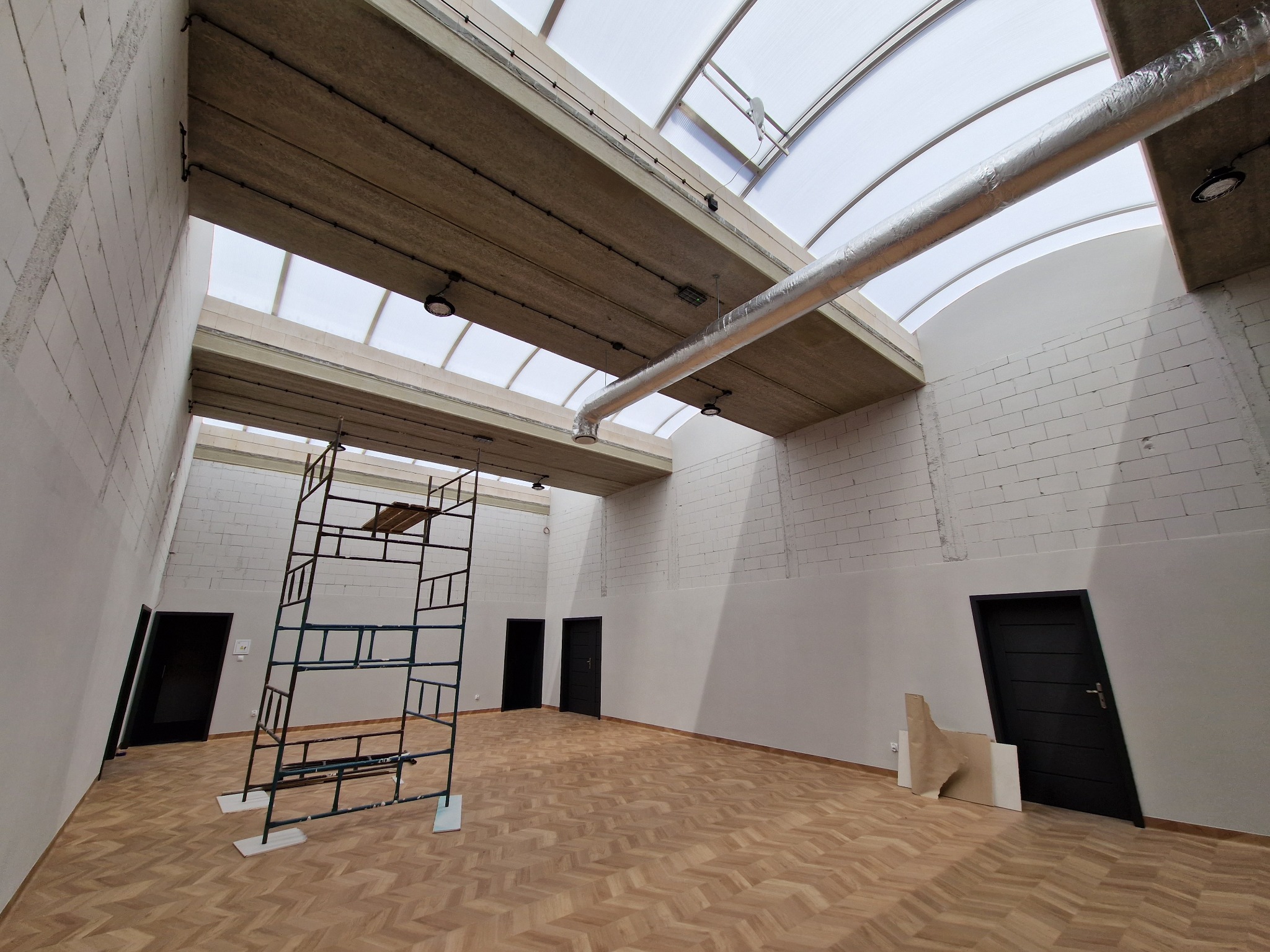Wnętrze hali w trakcie prac wykończeniowych: widoczne rusztowanie, instalacja wentylacyjna pod sufitem z świetlikami i częściowo pomalowane ściany z bloków.