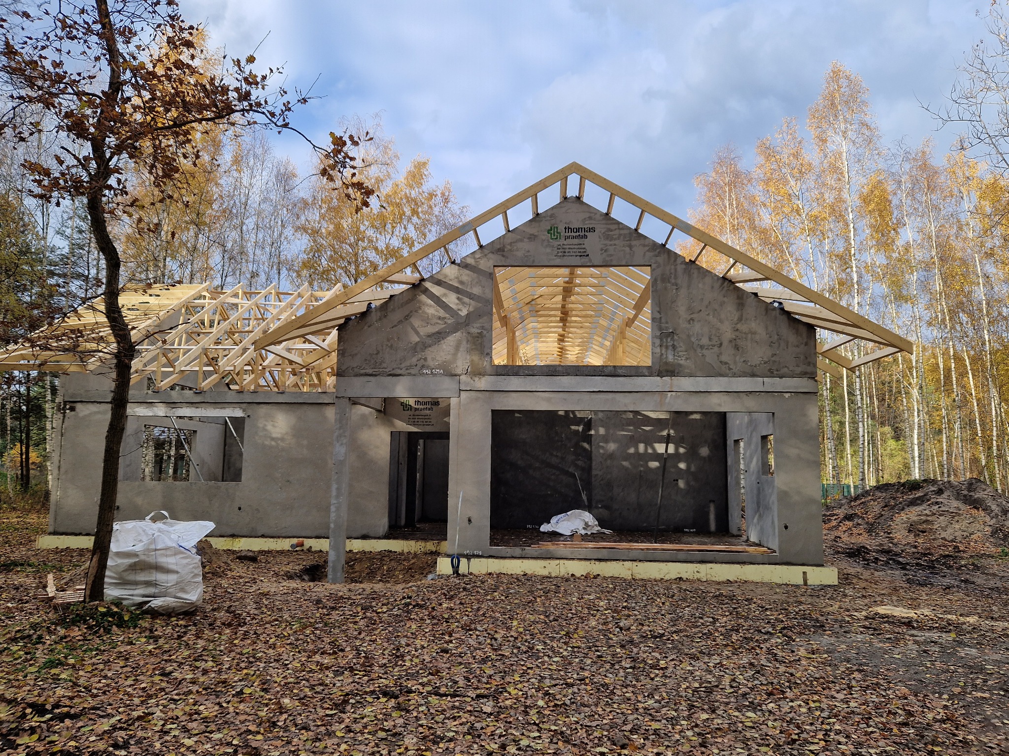 Surowy stan domu jednorodzinnego z drewnianą konstrukcją dachu w trakcie budowy, jesienne otoczenie, widoczny worek z materiałami budowlanymi na tle opadłych liści.