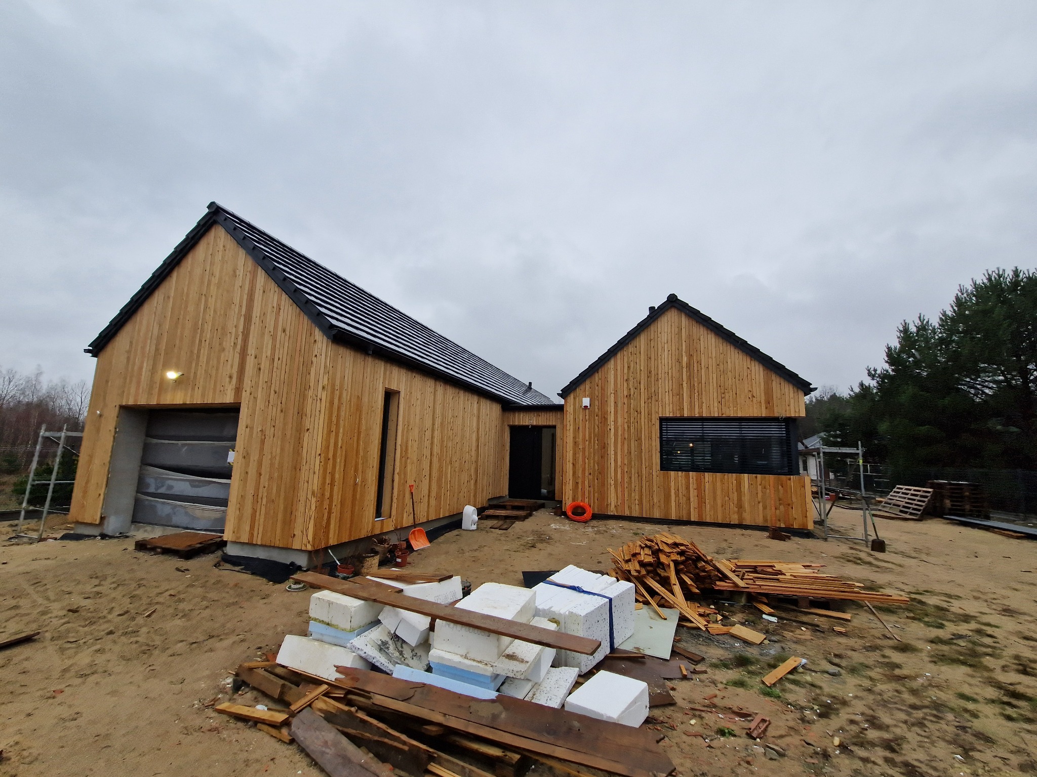 Nowoczesny dom z drewnianą elewacją w trakcie budowy, widoczne materiały budowlane i drabina. Pochmurne niebo w tle.