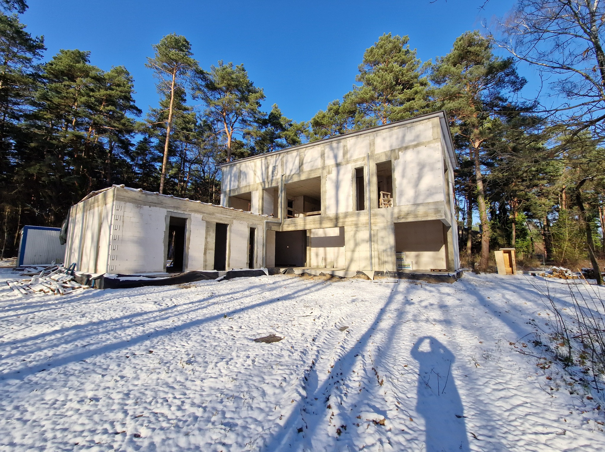 Surowy stan dwukondygnacyjnego domu z betonu komórkowego w zimowej scenerii, otoczonego lasem iglastym i śniegiem. Widoczne ślady opon i cień fotografa.