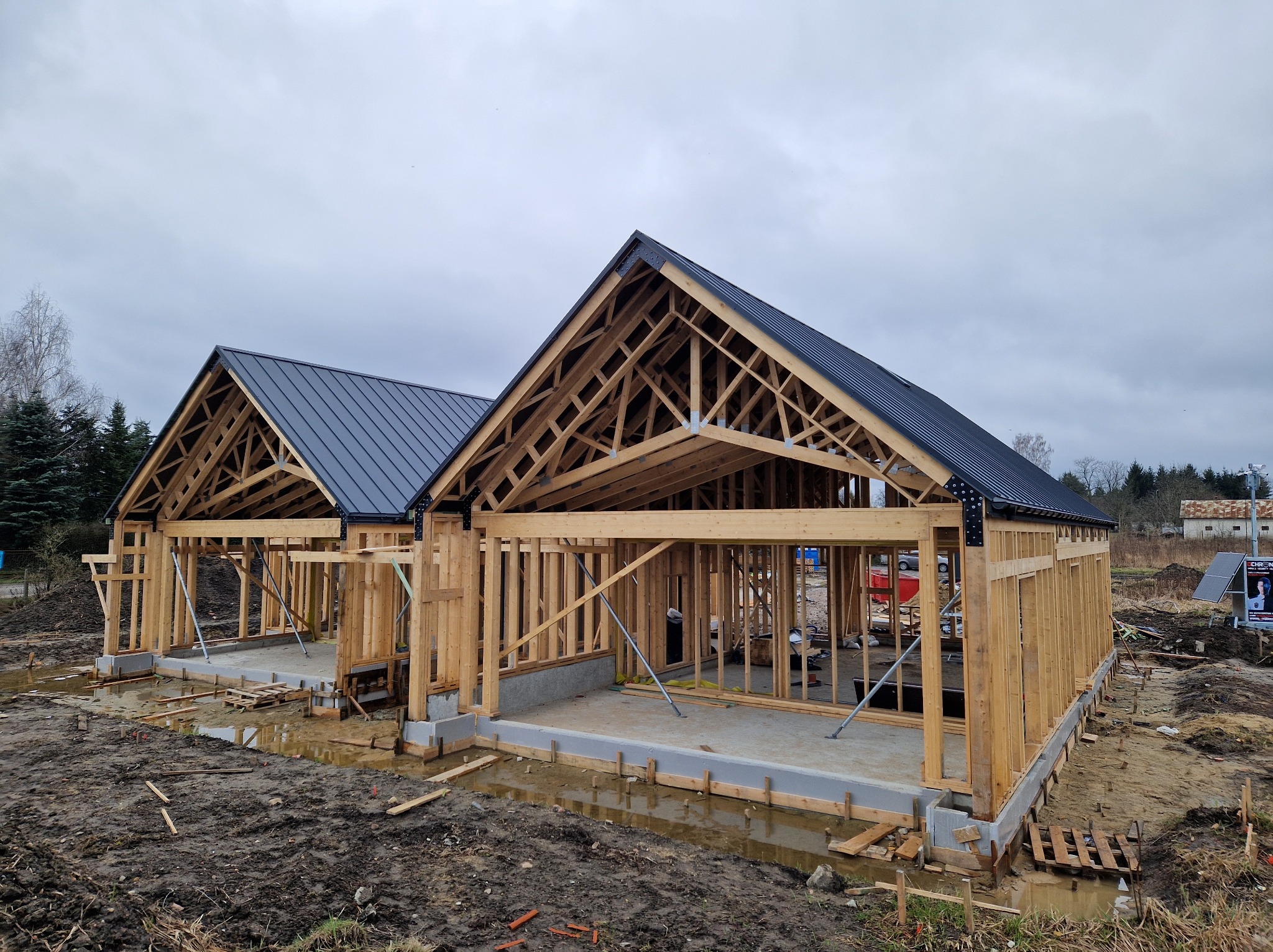 Dwa domy w stanie surowym otwartym z drewnianą konstrukcją szkieletową i częściowo zamontowanym dachem z blachy na tle pochmurnego nieba.