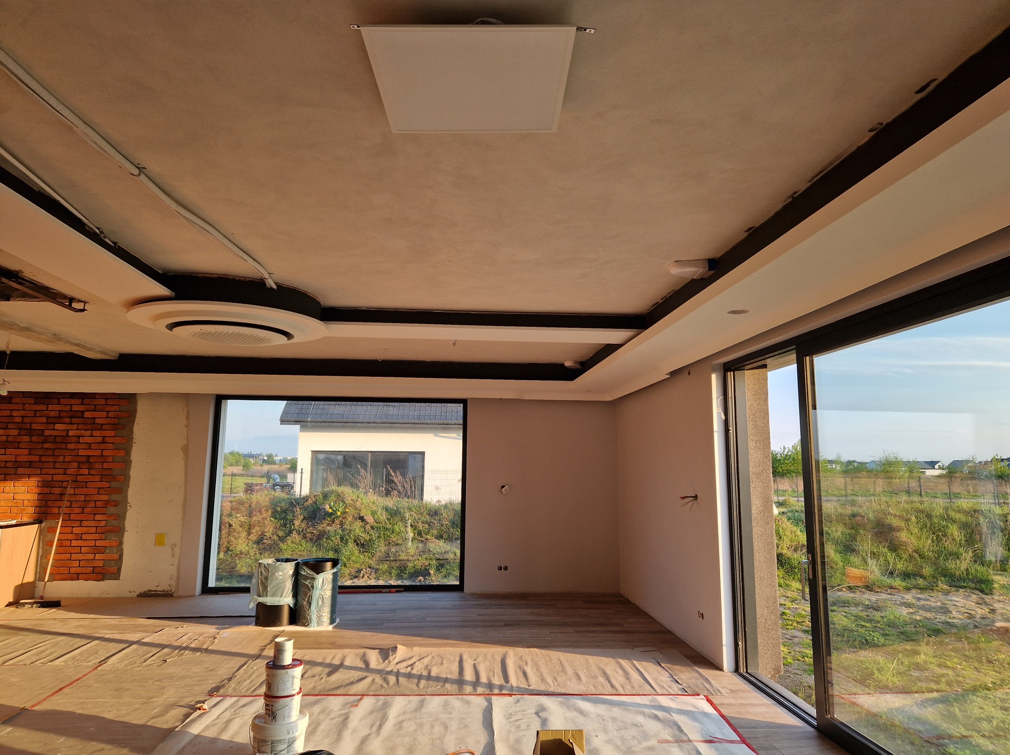 Wnętrze domu w trakcie wykańczania: widoczne okna, ściany przygotowane do malowania, podłoga zabezpieczona folią, elementy instalacji sufitowej.