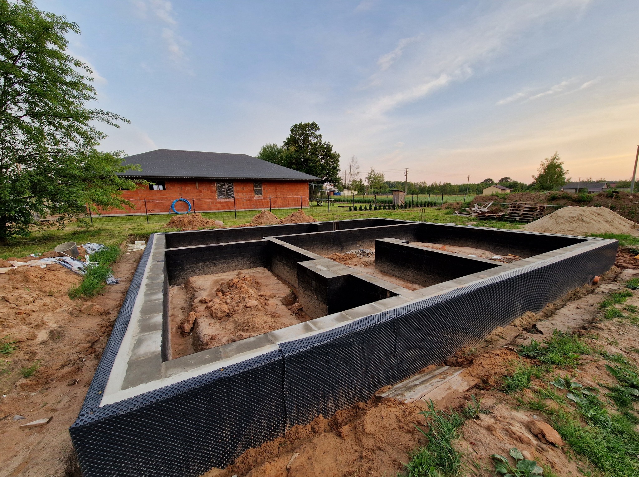 Fundamenty domu jednorodzinnego z izolacją, wykonane w Raszynie. Widok na budynek w tle, teren budowy w naturalnym oświetleniu, z perspektywy lekko z góry.