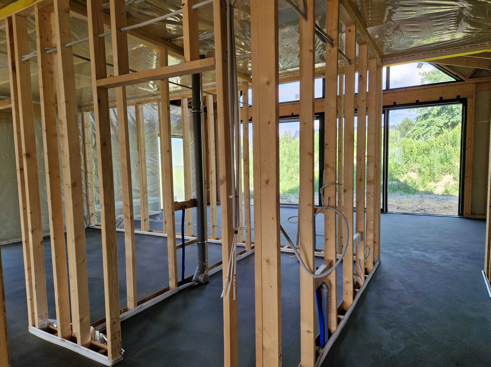 Wnętrze domu w trakcie budowy: drewniana konstrukcja szkieletowa, instalacje wod-kan i elektryczne w ścianach, widok na zieleń za oknem.