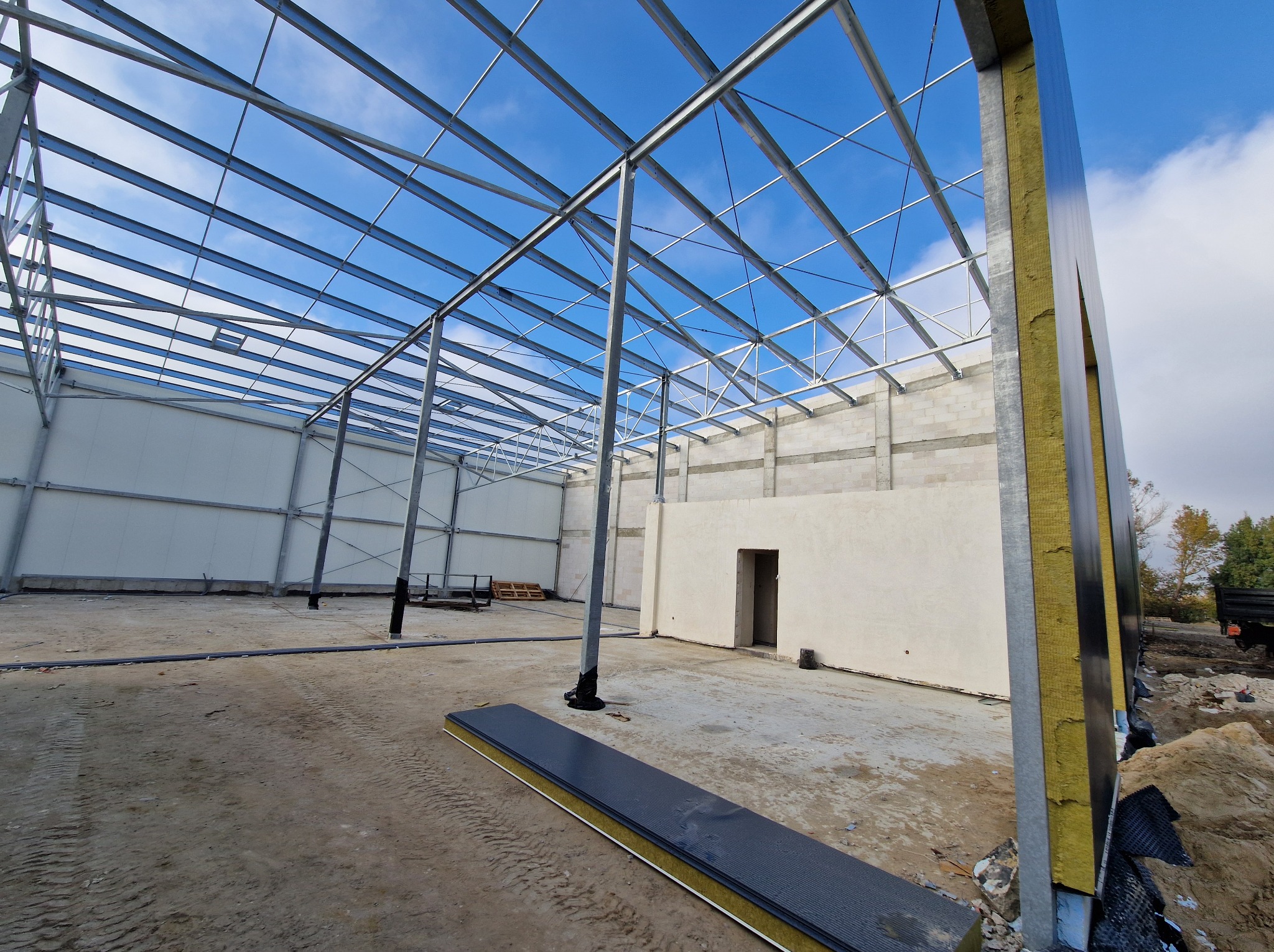 Wnętrze hali w budowie: stalowa konstrukcja dachu, ściany z betonu komórkowego i płyty warstwowe. Widoczne elementy izolacji. Jasne niebo w tle.
