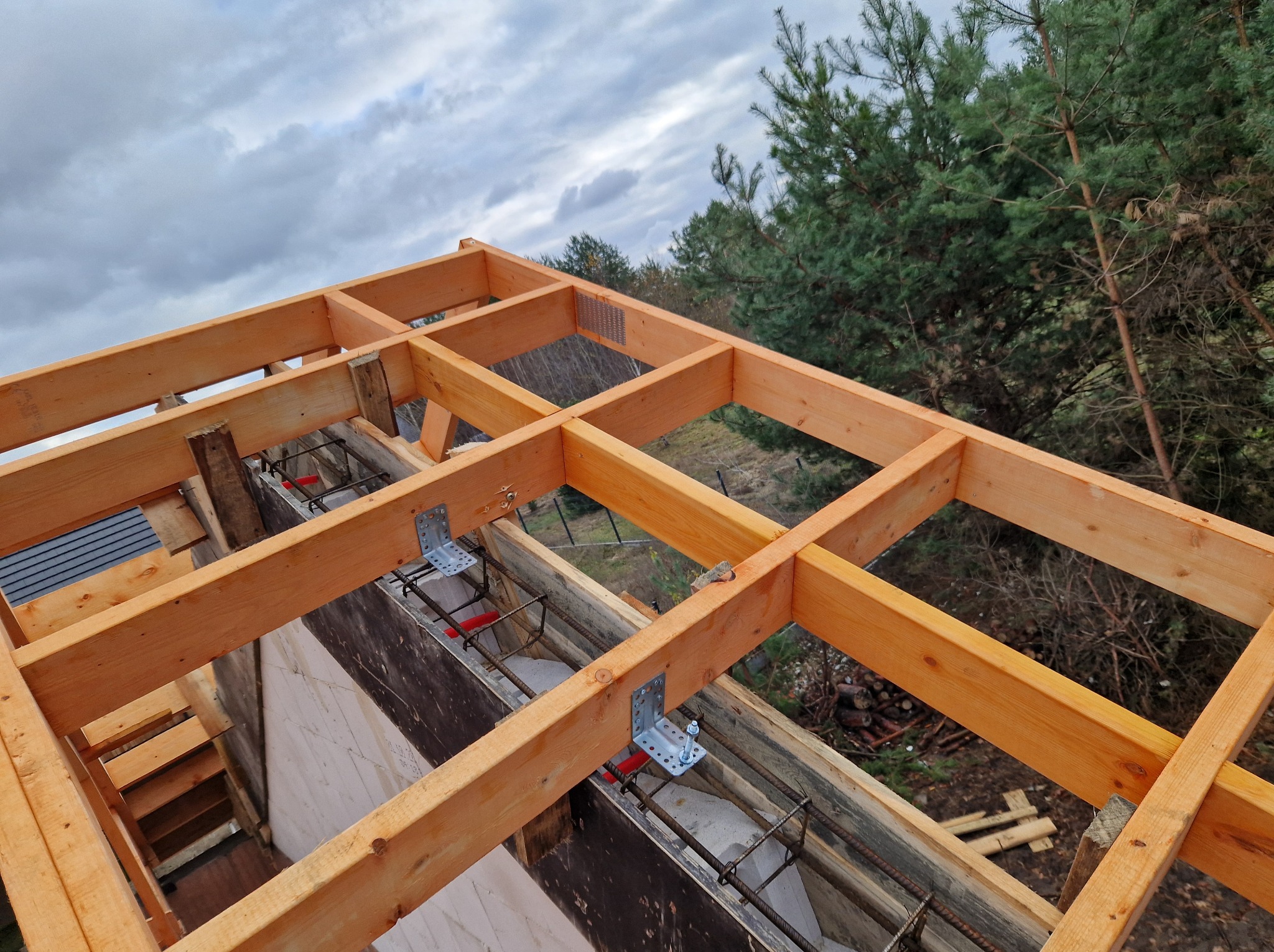 Drewniana konstrukcja szalunku stropu z widocznym zbrojeniem i łącznikami metalowymi, na tle pochmurnego nieba i zieleni drzew. Widok z góry na plac budowy.